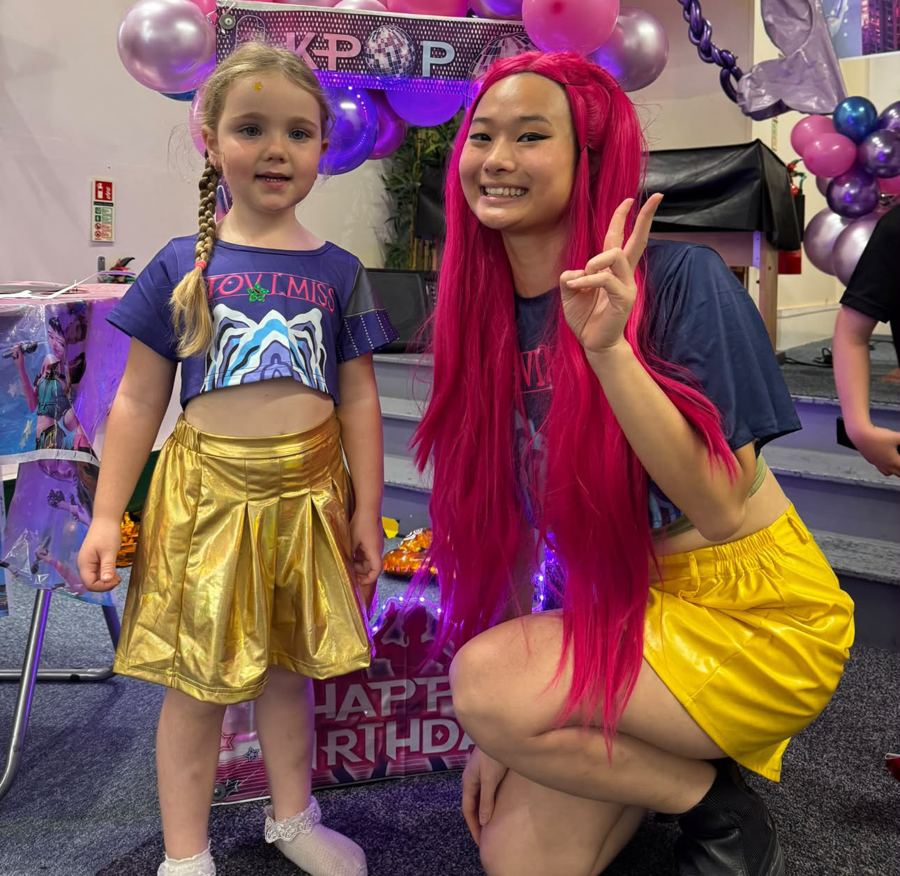 Performer in a pink wig posing with a girl in a gold skirt in front of K-pop balloons